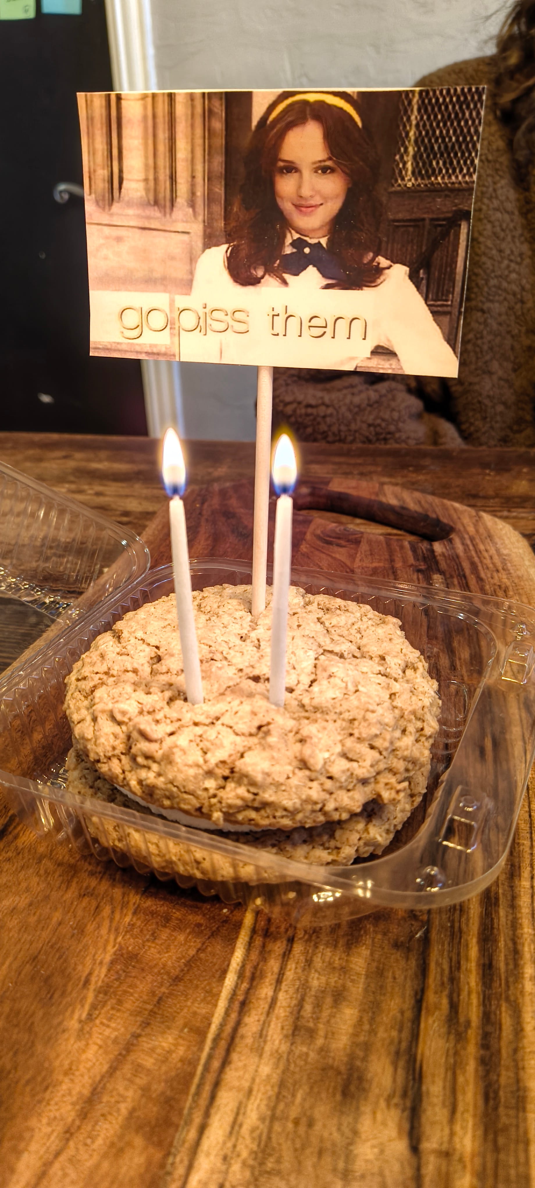 A large oatmeal cookie whoopie pie with two candles and a small sign riffing on the Gossip Girl "Go Piss Girl" meme reading "Go Piss Them"