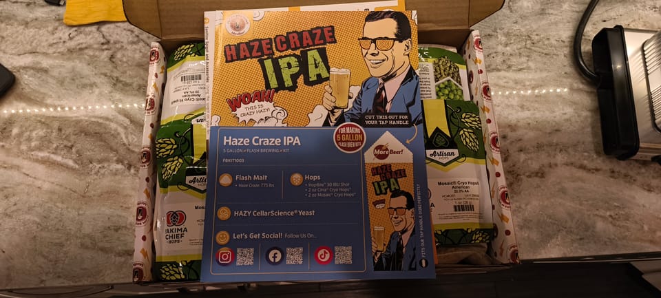 Photo of a beer kit on a marble counter. The kit is the "Haze Craze IPA" from MoreBeer.com, showing an illustration of a white man in a suit and tie, holding a hazy IPA in his right hand